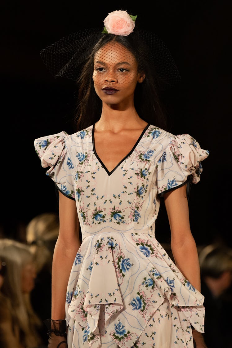 A female model walking a runway in a white dress with blue flower paintings and with a black veil wi...