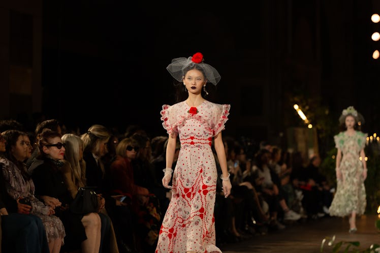 A female model walking while wearing a white and red gown and a white veil with a rose on her head