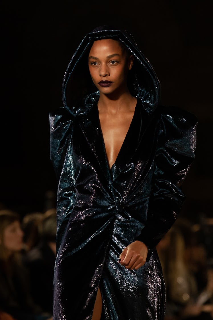 A female model walking a runway while wearing a black sequined coat with a cowl