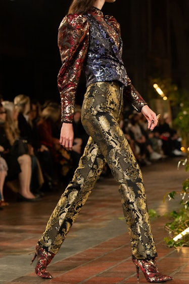 A female model walking a runway while wearing golden sequined pants and a blue and red sequined shir...
