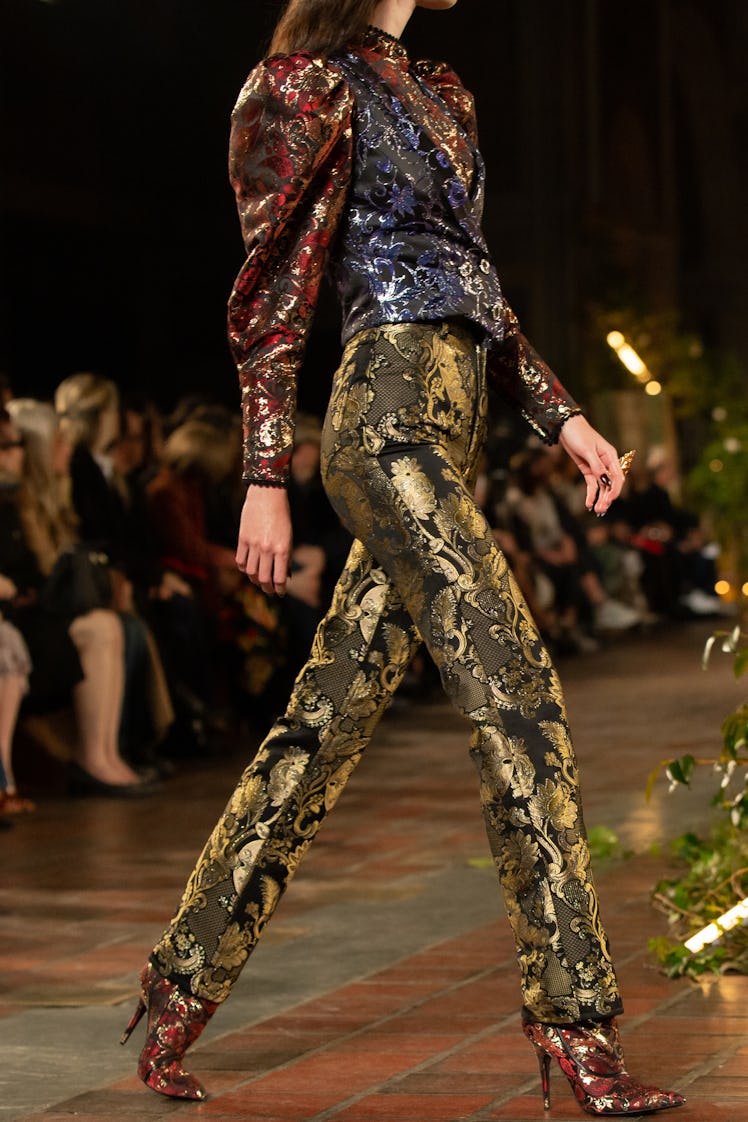 A female model walking a runway while wearing golden sequined pants and a blue and red sequined shir...
