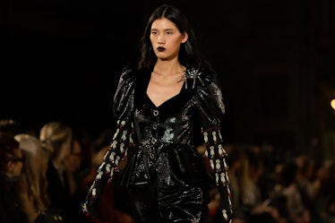 A female model walking a runway while wearing a black latex blazer over a black shirt 