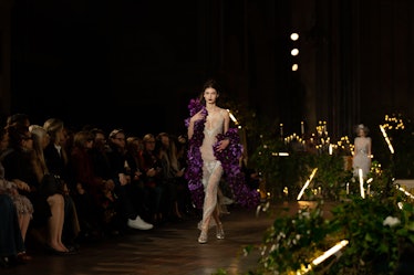 A female model walking a runway in a white see-through dress paired with a purple robe 
