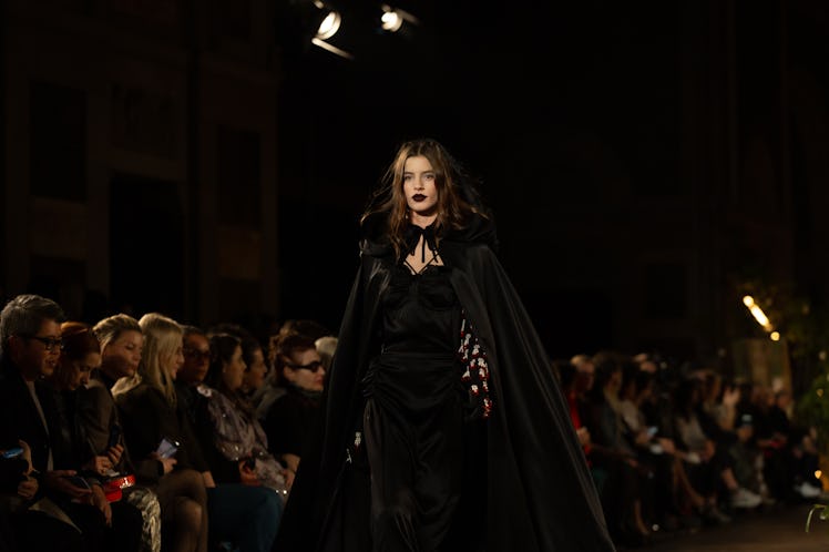 A female model walking a runway while wearing a black dress and a black mantle