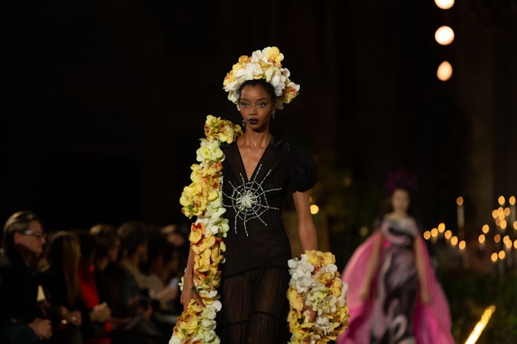 A female model walking with a yellow robe while wearing a black dress with a white spider net drawin...
