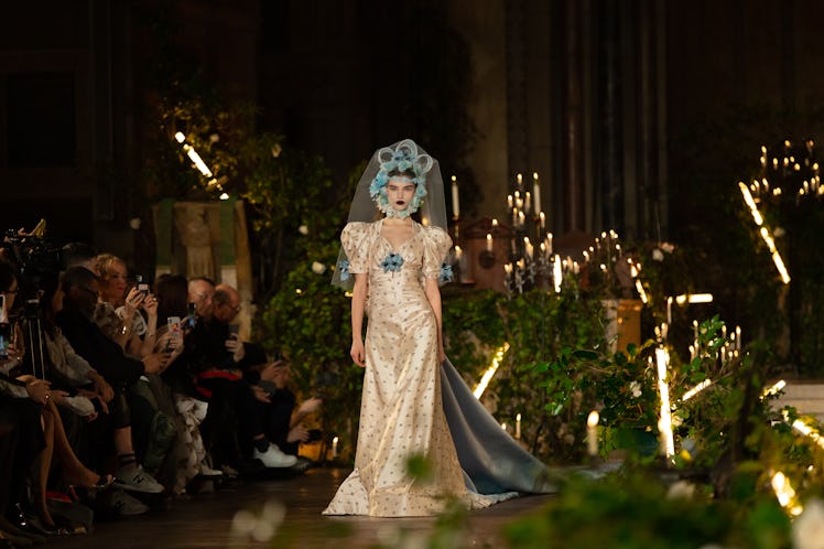 A female model walking a runway while wearing a white gown and a light blue veil