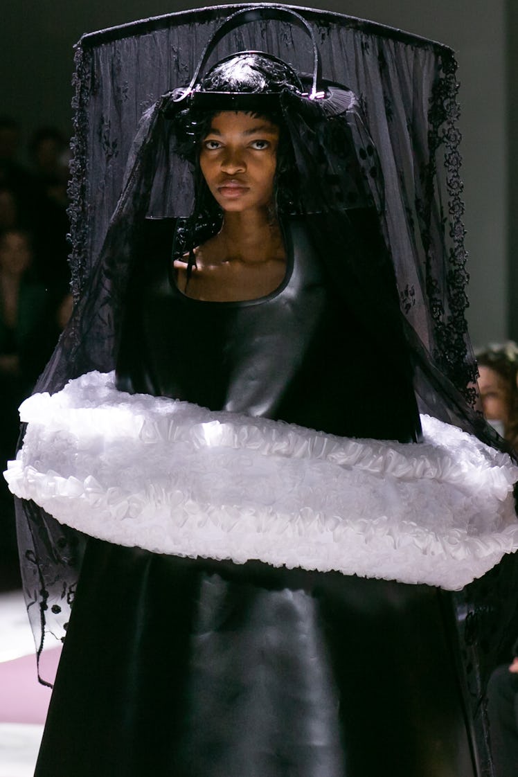 A model walking the Comme Des Garcons runway in a black gown and headpiece, and a white soft accesso...