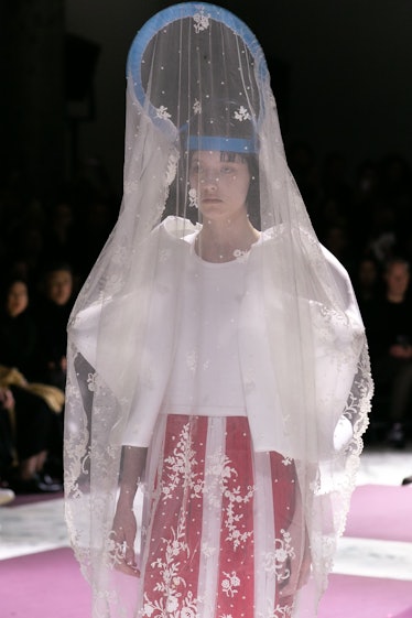 A model walking the runway in a white top with puffy sleeves, a red skirt and a blue headpiece with ...