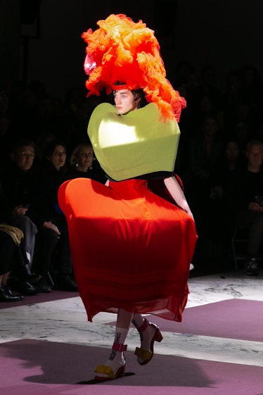 A model in a green box-like top, an orange box-like skirt, and an orange headpiece at Comme Des Garc...