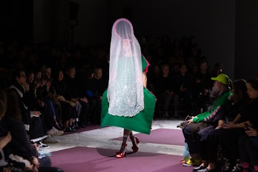 A model on the Comme Des Garcons runway in a green dress, red heels, and a pink with black headpiece...
