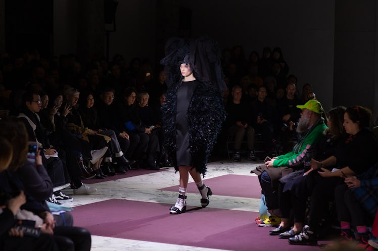 A model in Comme Des Garcons' show in a black dress, black shoes, a feathered black headpiece and ma...
