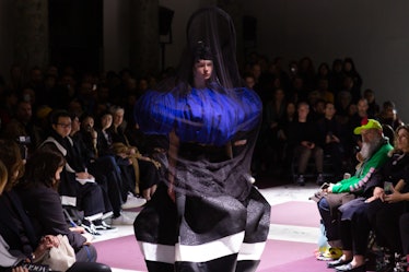 A model on the Comme Des Garcons runway in a black dress with a white stripe and blue feathers with ...