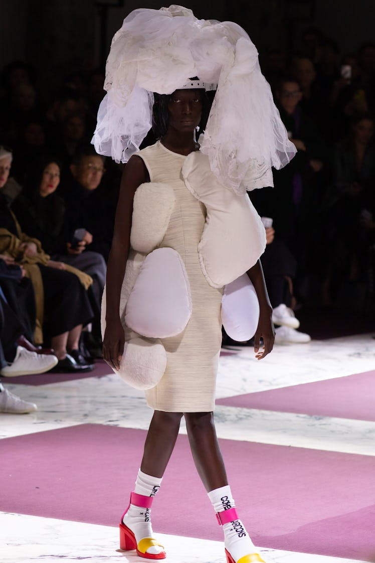 A model on the Comme Des Garcons runway in a white dress with accessories resembling pillows and a l...