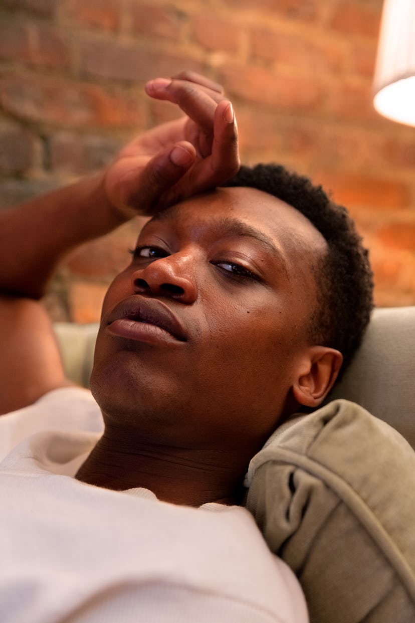 Ato Blankson-Wood lying on a couch with a hand on his forehead while wearing a white shirt