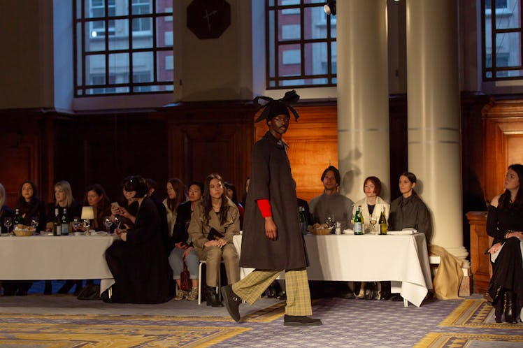 A model in a black coat and green trousers and a black hat at Molly Goddard’s London Fashion Week Sh...