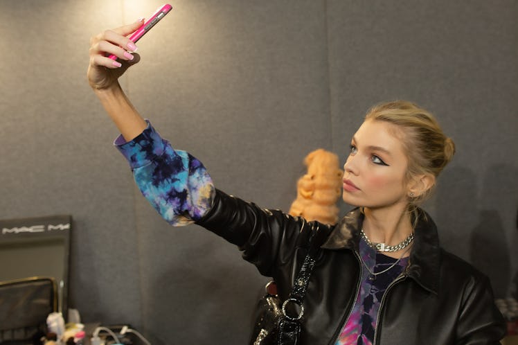 A model in a blue tie-dye sweater and a black jacket taking a selfie backstage at the Moschino Fall ...