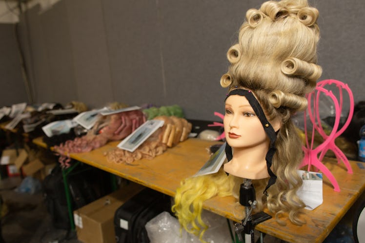 A doll head with a blonde wig placed on a wooden desk with other wigs backstage at the Moschino Fall...
