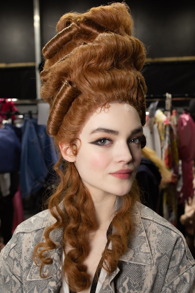 A portrait of a model with a large brown wig in a grey jacket backstage at the Moschino Fall 2020 sh...