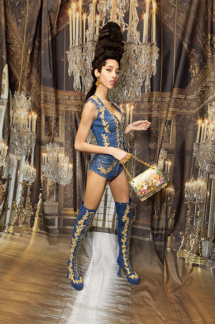 A model in a blue-gold short playsuit and matching lace-up boots backstage at the Moschino Fall 2020...