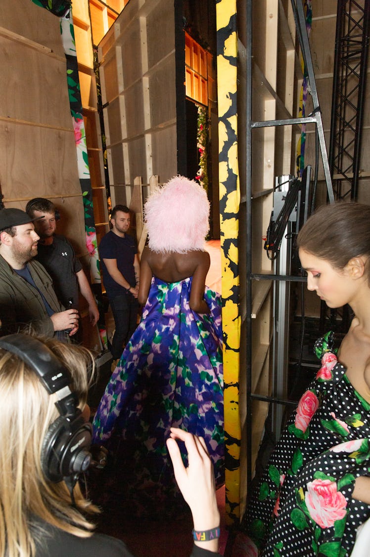 A model in a blue-pink floral dress and pink feather hat surrounded by people backstage at Richard Q...