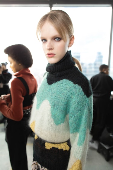 A blonde female model posing for a photo while wearing a white, light blue, and black sweater 
