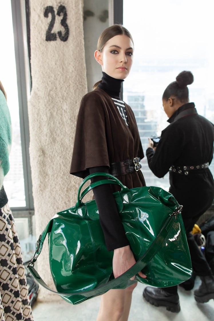 A female model standing while wearing a dark brown dress and a green bag
