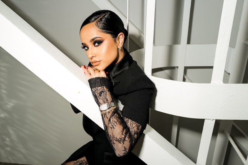 Becky G posing in a black lace dress and leaning on a white building construction element