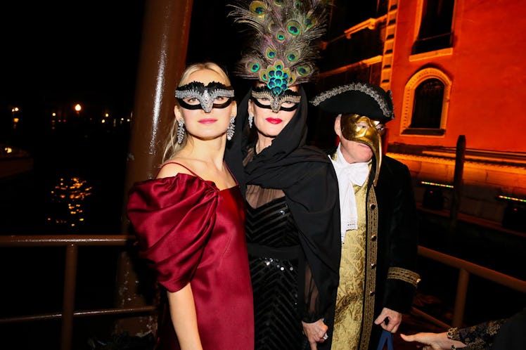 A woman in a red dress and black mask and a woman in a black dress and grey feather mask at the Save...