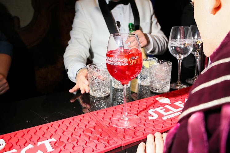 Glass with red liquor on a bar table top inside the Save Venice party and a man in a white tuxedo