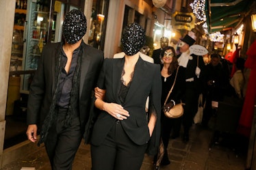 A man and a woman in black suits walking down a street in Venice wearing masks for the Save Venice p...