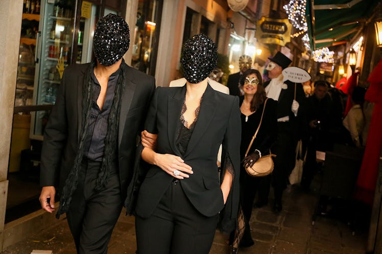 A man and a woman in black suits walking down a street in Venice wearing masks for the Save Venice p...