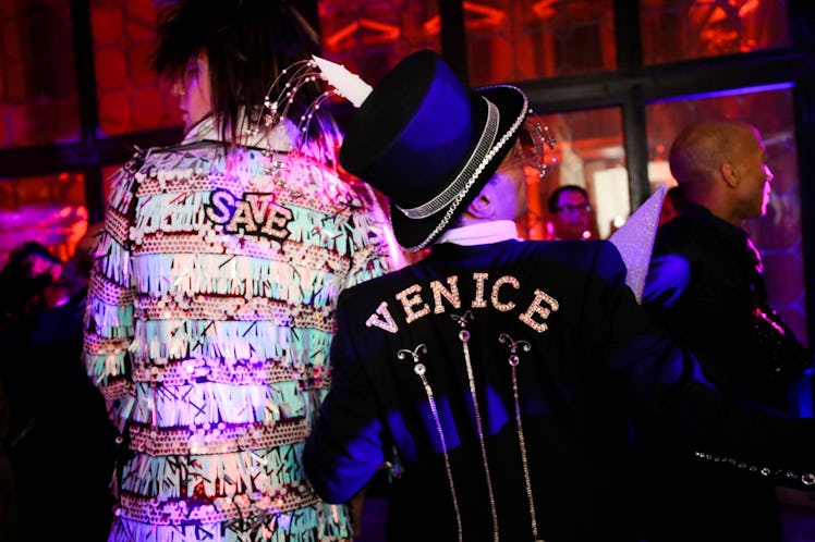 A man in a white-beige pastel blazer and the text 'SAVE' on his back and a man in a black blazer wit...