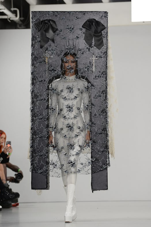 A model with a frame attached to her shoulders, with black lace draping from it, covering her face w...