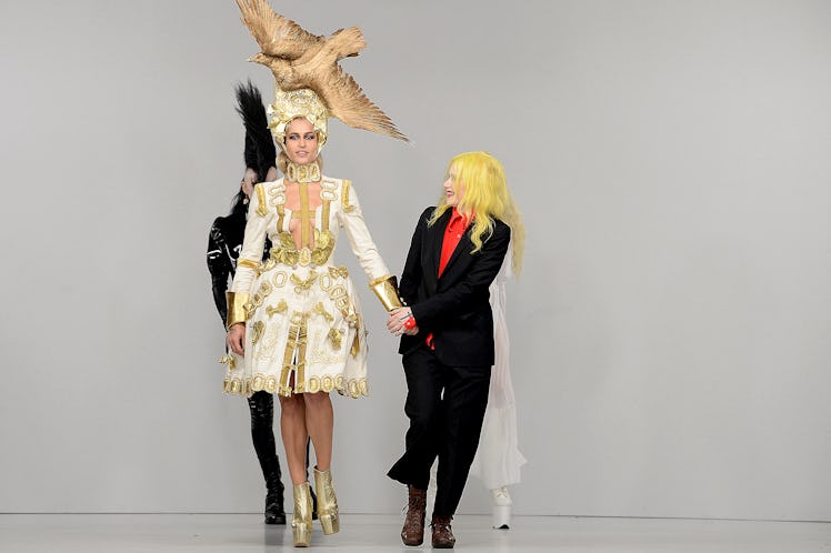 A model in a white mini dress with an enormous gilded taxidermy bird atop her head walking and holdi...