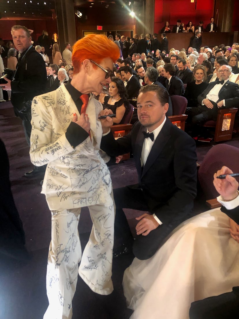 Sandy Powell in a white suit with black signatures next to Leonardo di Caprio in a black suit, white...