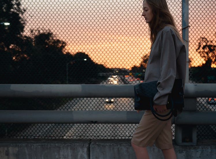 A woman walking while wearing a grey shirt and brown shorts and carrying a black bag