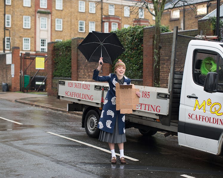 A girl holding a sign that says "Welcome to London", while wearing Prada coat, top, skirt, and shoes