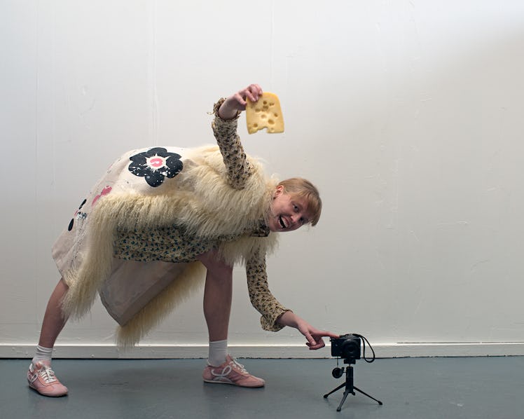A model holding a piece of cheese,  bending over towards the floor, while wearing a Miu Miu gilet