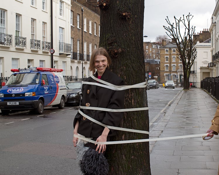 A model wearing a Miu Miu jacket, AS Colour T-shirt, and a Sacai skirt, while being taped to a tree
