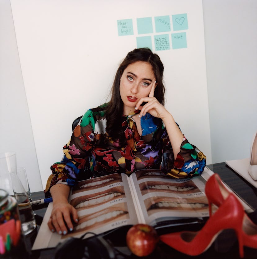 Catherine Cohen in a floral top posing in with an opened book in front of her