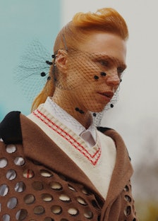 A model at Paris Fashion week in a beige sweater, brown coat, and a black veil-like net on her face