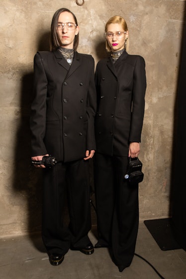Two models wearing black suits by Marine Serre, while standing and posing backstage