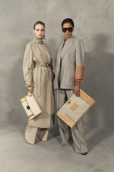 A model in a beige coat and a model in a grey suit both holding beige bags backstage at the Givenchy...