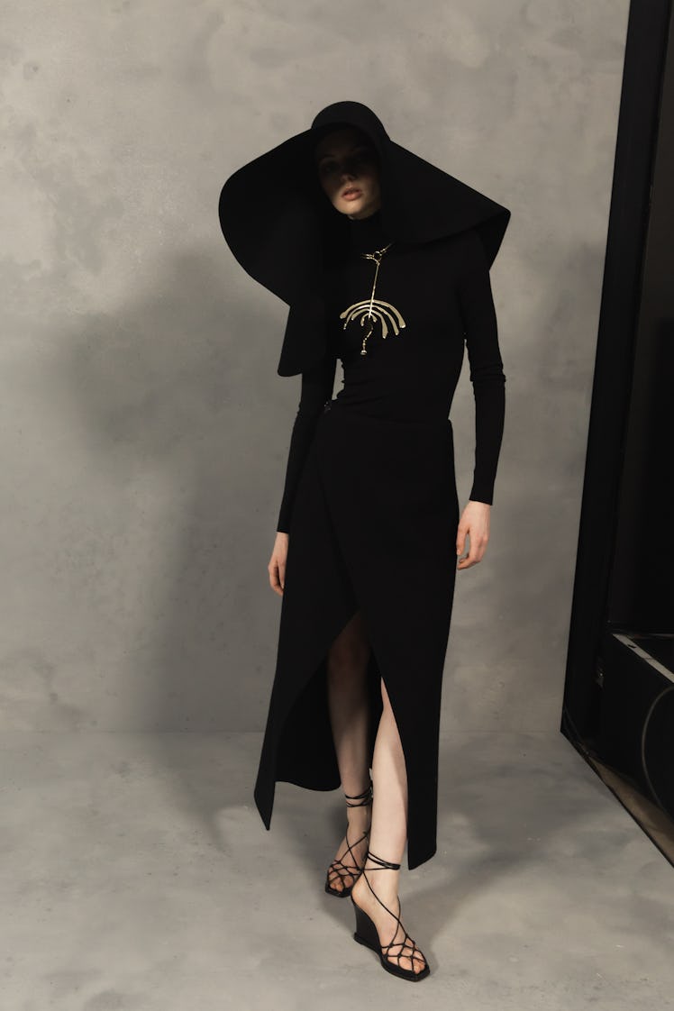 A model wearing a black coat, black sandals and an oversized black hat backstage at the Givenchy Fal...