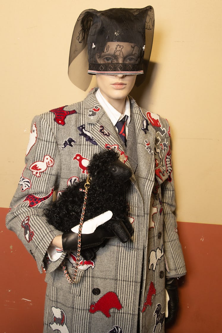 A model at Thom Browne's show in a plaid jacket with animals on it and a black lace headpiece, holdi...