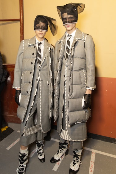 Two models at Thom Browne's show in long grey puffer jackets, black and white shoes, and a black lac...
