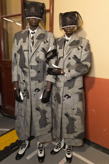 Two models backstage at Thom Browne's show in long grey camo jackets, black and white shoes and blac...