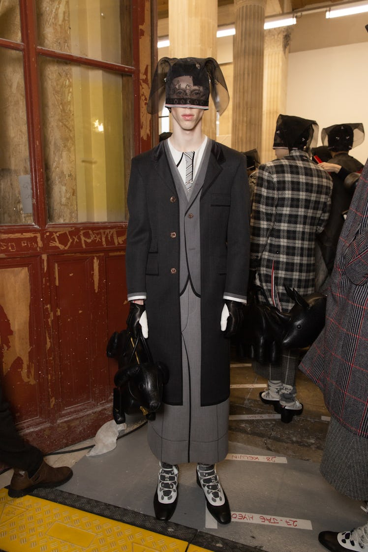 A model backstage at Thom Browne's fall 2020 show in a grey suit, black jacket, animal-shaped bag an...