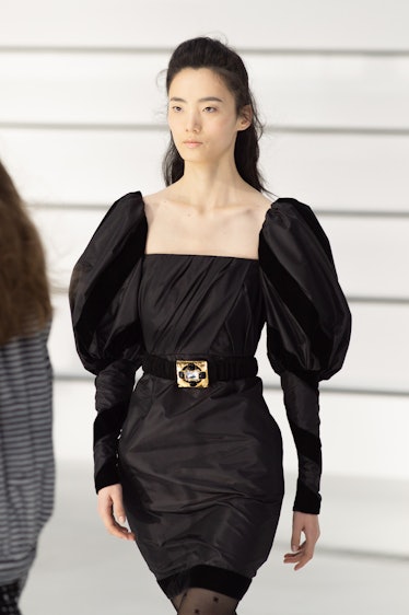 A model walking in a black dress with gold detail on belt at the Chanel fall 2020 show