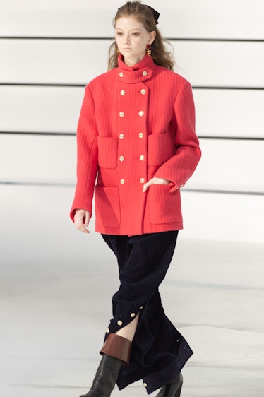 A model walking in an orange jacket, a black skirt and brown boots at the Chanel fall 2020 show